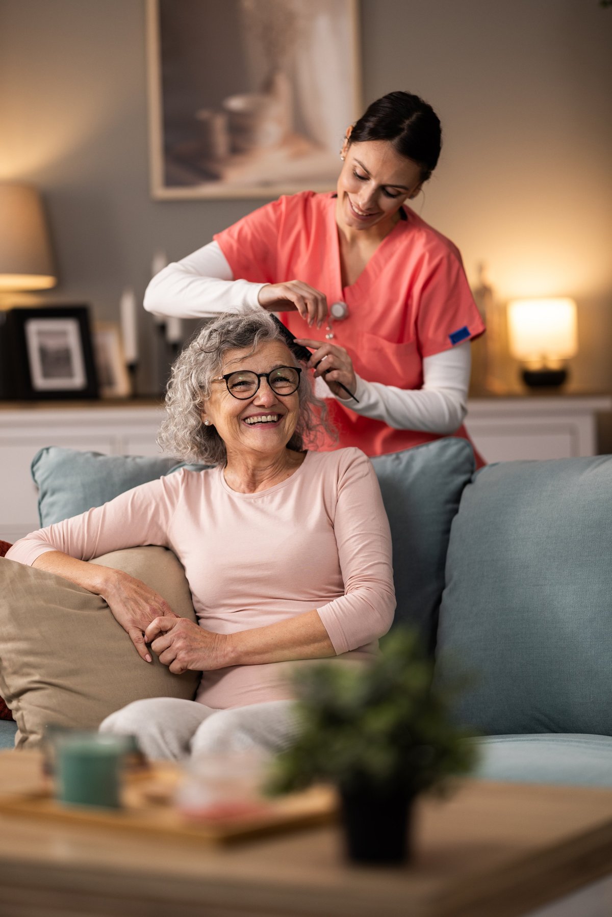 Young cheerful nurse combing senior woman's hair while she is sitting on couch in living room, talking and smiling